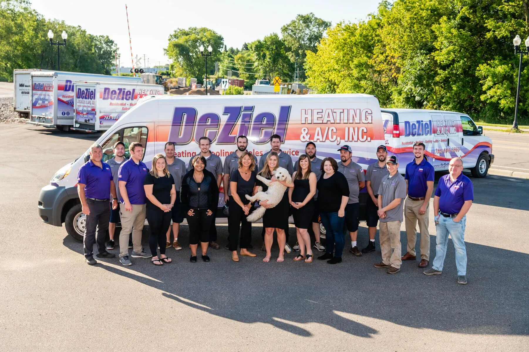 A Group Of People Stands In Front Of A Deziel Heating &Amp; A/C, Inc. Van In A Parking Lot, Some Proudly Wearing Company Uniforms, With A Dog Happily Seated In The Center. Multiple Company Vans Are In The Background, Hinting At Their Fall Offer For Air Exchanger Maintenance.